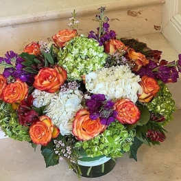 Low round arrangement of orange roses, green and white hydrangeas, and purple blooms in a glass vase