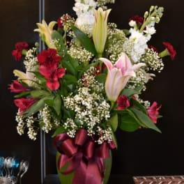 Tall bouquet of lilies, red flowers, and baby's breath in a vase with a ribbon
