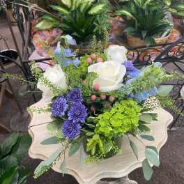 Low arrangement of white roses, purple mums, blue blooms, and green hydrangea in a vase on a small table