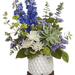 Tall blue delphinium, white mums, purple accents, and a succulent in a white ceramic pot on a wooden stand.