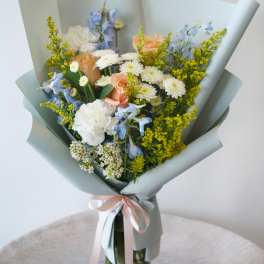 Bouquet of peach roses, white daisies, and blue flowers wrapped in pale paper in a glass vase