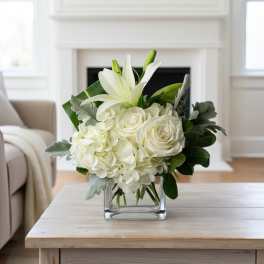 Low white arrangement of lilies, roses, and hydrangeas in a clear glass cube vase on a light wood table