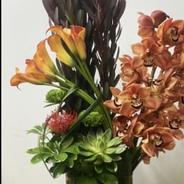Tall arrangement of orange calla lilies and orange orchids in a glass vase