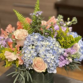Pastel bouquet with blue hydrangeas, peach roses, and lavender flowers in a glass vase