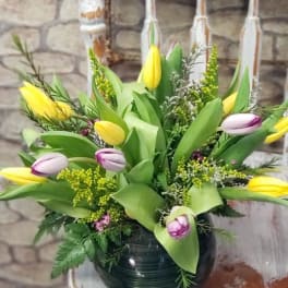 Low arrangement of yellow and purple-tipped tulips in a round glass vase on a rustic white chair