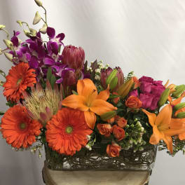 Bright mixed bouquet with orange lilies, gerbera daisies, and purple orchids in a glass vase