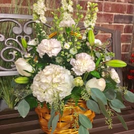 White floral arrangement in a wicker basket with lilies, hydrangea, and tall blooms