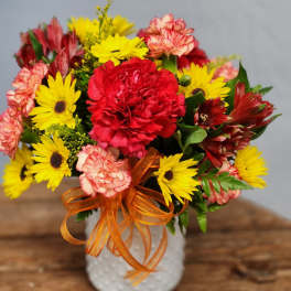 Bright arrangement of red and pink carnations with yellow daisies in a white vase with orange ribbon