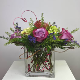 Pink and purple roses arranged in a clear square vase with curly wire accents