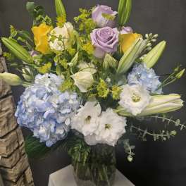 Mixed bouquet with blue hydrangeas, roses, lilies, and white blooms in a glass vase