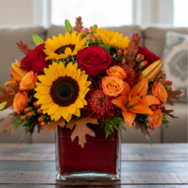 Autumn arrangement with sunflowers, red roses, and orange lilies in a square glass vase