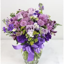 Lavender and purple mixed flower arrangement with roses and mums in a clear vase with a satin ribbon bow
