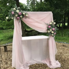 Pink draped wedding arch with floral arrangements and a table beneath it