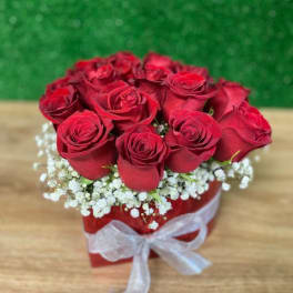 Short arrangement of red roses in a red box with white accent flowers and a white ribbon bow