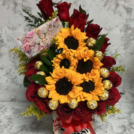 Arrangement of yellow sunflowers and red roses with gold-wrapped chocolates in a wooden box with a red bow and card.