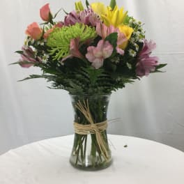 Mixed bouquet of yellow lilies, peach roses, and pink blooms in a clear vase with a raffia tie