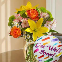Bouquet of yellow lilies, orange roses, and pink flowers in a glass vase with a Get Well Soon balloon.