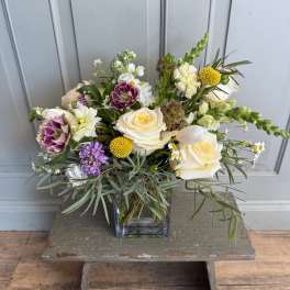 Mixed bouquet of cream roses, purple blooms, and yellow accents in a glass vase