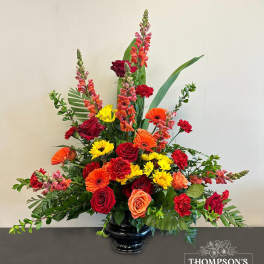Tall mixed arrangement of red roses, orange gerbera daisies, yellow mums and snapdragons in a black vase.