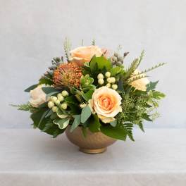 Peach roses arranged in a low gold vase with mixed greenery