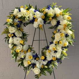 Heart-shaped floral wreath with white roses, yellow blooms, and blue accents on a stand