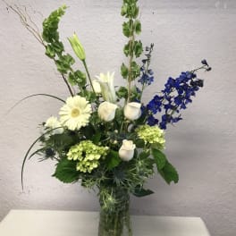 Tall floral arrangement with white roses, lilies, and blue flowers in a glass vase