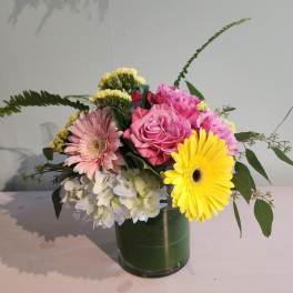 Mixed bouquet of pink roses, gerbera daisies, hydrangea, and yellow gerbera in a leaf-lined glass vase