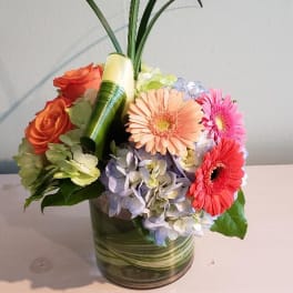 Compact arrangement of bright gerbera daisies, roses, and hydrangeas in a short glass vase with tall looping grass.
