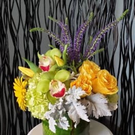 Compact yellow and green floral arrangement with orchids, roses, hydrangea and purple accents in a glass vase