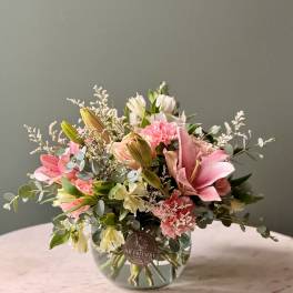 Low glass vase of pink lilies, carnations, and cream alstroemeria arranged in a rounded bouquet.