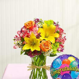 Bouquet of yellow lilies and orange roses in a glass vase beside a congratulatory balloon