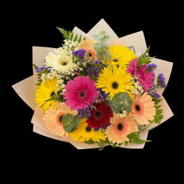 Hand-tied bouquet of multicolored gerbera daisies with white and purple accents wrapped in pale paper