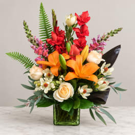 Bright mixed bouquet in a green glass vase with orange, red, cream, and pink blooms.