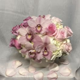 Low arrangement of pink orchids, roses, and white hydrangeas in a white cube vase with scattered petals.