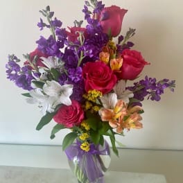 Tall mixed bouquet of pink roses, purple stock, and white and peach alstroemeria in a glass vase.