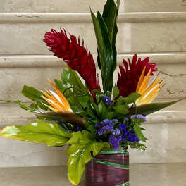 Tall tropical arrangement with red ginger, orange blooms, and purple flowers in a striped square glass vase.