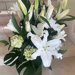 Tall arrangement of white lilies and hydrangeas with monstera leaves in a clear glass vase