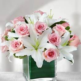 Pink roses and white lilies in a clear glass cube vase lined with a green leaf.