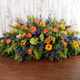 Low spray arrangement of orange roses, yellow daisies, and purple flowers in an elongated shape