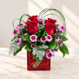 Red roses and pink daisies in a red square vase with looping greenery