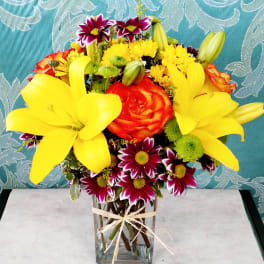 Bright bouquet of yellow lilies, orange roses, and mixed daisies in a clear rectangular vase