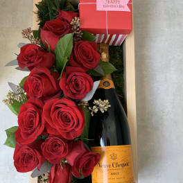 Red roses arranged with a champagne bottle and gift box in a wooden tray