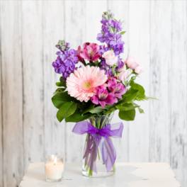 Mixed bouquet of pink and purple flowers in a clear vase with purple bow beside a lit candle.