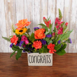 Bright mixed arrangement of orange, pink, purple, and yellow flowers in a rectangular Congrats container