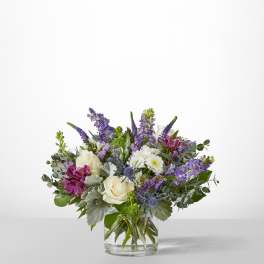 Bouquet of white roses, purple flowers, and blue thistle in a clear glass vase