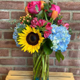 Bouquet of sunflowers, pink roses, blue hydrangeas, and orange tulips in a glass vase