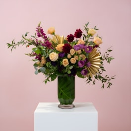 Tall peach, burgundy, and purple flower arrangement with gold accents in a green-filled glass vase.