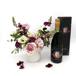 Pink and white floral arrangement in a white vase with a bottle of sparkling wine and gift box.