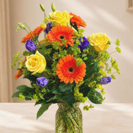 Bouquet of yellow roses, orange gerbera daisies, and purple blooms in a glass vase
