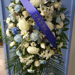 Standing funeral spray of white and blue flowers with a purple ribbon reading DADDY
